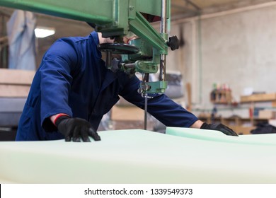 Process of furniture production. A young man in a dark coat cut the foam for the sofa on a cutting machine. Behind him there are a lot of finished sofas. He is wearing a safety glasses and gloves. - Powered by Shutterstock