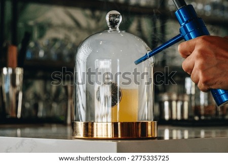 Process of infusing cocktail with smoke using cloche. Technical shot of smoke being pumped into a glass cloche covering a cocktail on a white marble counter.