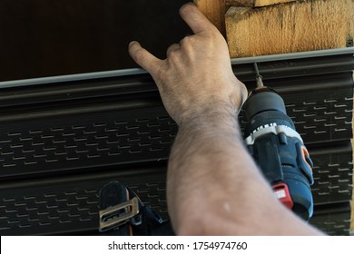 The Process Of Installing Vinyl Siding On A Wooden Beam. The Screwdriver Tightens The Screw. Hand Holds A Plastic Sheet.