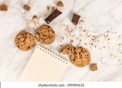 The Process Of Making Chocolate Chips Cookies. Overhead Shot Of Biscuits With Chocolate Pieces, Flour, And Cane Sugar Around Them, With A Blank Notepad For Copy Space