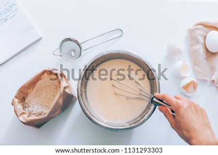 Process of making the dough, woman's hand whips eggs and flour in bowl next to the handwritten recipe and sieve, top view. Flat lay composition of ingredients and batter.