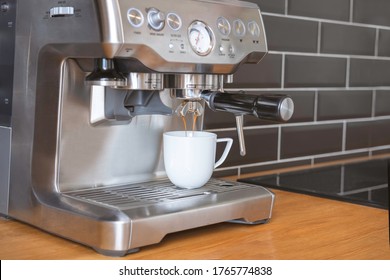 The process of making espresso coffee in a coffee machine (maker) with white cup in the kitchen with a black wall and a wooden worktop at home.