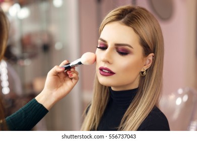 Process of making makeup. Make-up artist working with brush on model face. Portrait of young blonde woman in beauty saloon interior. Applying tone to skin.