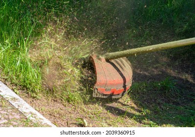 The Process Of Mowing A Lawn With A Hand Mower. A Worker Mows Tall Grass With An Electric Or Gasoline Trimmer In A City Park Or Backyard. Garden Care Tools And Equipment.