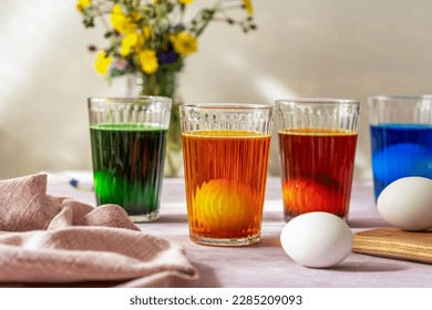 Process of painting Easter eggs. Four glasses with different colours, eggs inside with dye liquid, napkin, wild flowers