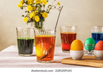 Process of painting Easter eggs. Four glasses with different colours, eggs inside and already painted, wild flowers
