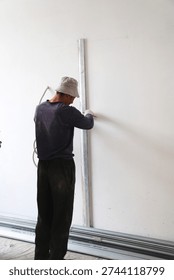 The process of preparing for partition installation or wall leveling at a construction site. A worker wearing a bucket hat and protective gloves stands with his back to the camera and holds a long aluminum construction straight edge against a white wall while marking with a red pencil. Metal guide profiles for drywall lie on the floor. The shot clearly demonstrates the initial stage of finishing works where high precision and adherence to room geometry are essential. Perfect for DIY renovation content construction crew services and architectural design