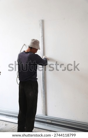 The process of preparing for partition installation or wall leveling at a construction site. A worker wearing a bucket hat and protective gloves stands with his back to the camera and holds a long aluminum construction straight edge against a white wall while marking with a red pencil. Metal guide profiles for drywall lie on the floor. The shot clearly demonstrates the initial stage of finishing works where high precision and adherence to room geometry are essential. Perfect for DIY renovation content construction crew services and architectural design