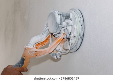 The process of sanding a concrete wall using a specialized electric sander during indoor renovation works. The tool is covered in white drywall dust, indicating an active phase of surface preparation.