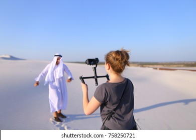 Process of shooting Arab guy on camera. Young woman holds camera in hands and guides Arab to stately man who poses and smiles, dances in middle of bottomless desert on hot summer day. Emirate with