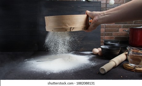 The process of sifting flour. Baking in a stylish kitchen. Kitchen utensils. A large wooden sieve. Snow-white flour on a black background - Shutterstock ID 1882088131