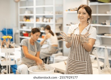 In process of working in pottery workshop, girl takes break, stands and rests, waves hello with her hands stained with clay. She shows resulting blank, future vase - Powered by Shutterstock