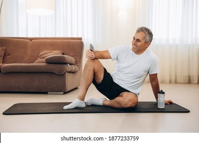 Procrastination During Fitness. Senior Man Resting After Home Workout With Water And Phone. Sporty Man After Practicing Yoga, Break In Doing Exercise, Relaxing On Yoga Mat, Texting On Smartphone.