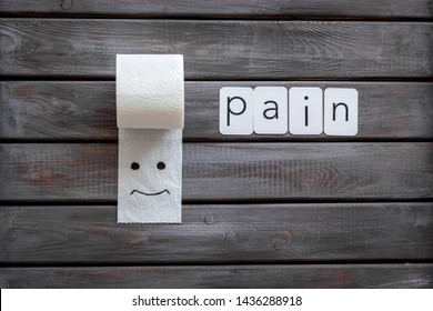 Proctology concept with pain text, toilet paper roll and painted face on wooden background top view
