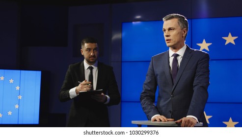 Producer And News Anchor In A Production Team In A News Studio Issuing Instructions While Standing In Front Of The EU Flag