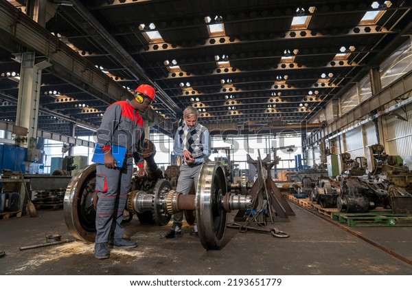 Production\
Manager Talking To African American Worker Next To Train Wheels At\
Train Factory. Area For Maintenance, Repair And Service Of Trains\
And Wagons. Quality Control In\
Manufacturing.