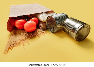 Products In A Craft Bag On A Yellow Background. Macaroni, Tomatoes, Rice And Canned Food Spilled Out Of The Bag Onto A Yellow Surface In Close-up. Food Delivery Concept.