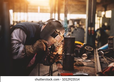 Profesional Welder In Protective Uniform And Mask Welding Metal Pipe On The Industrial Table With Other Tools In The Industrial Workshop.