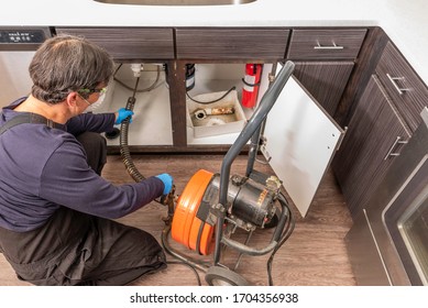 A Professinal Plumber Unclogging A Kitchen Sink Drain Using A Commercial Snake Auger, Wearing A Safety Mask Per Covid Guidelines From The US Government