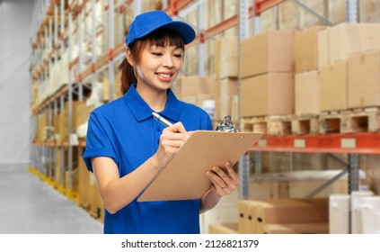 profession, job and people concept - happy smiling delivery woman in blue uniform with clipboard and pen over warehouse background