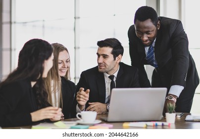 Professional adult African businessman leader pointing and consulting planning strategy with concentrated caucasian colleagues that gathered in front of laptop while sitting at office working table - Shutterstock ID 2044642166