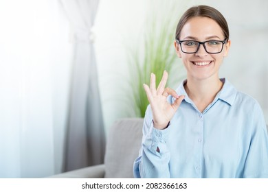 Professional advice. Expert approval. Good job. Satisfied cheerful smiling smart business woman in eyeglasses accepting with okay gesture on couch at light home interior.