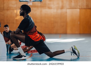 Professional african american basketball player is stretching his leg on training. Side view of a multicultural basketball player warming up on his training. In a blurry background are his teammates. - Powered by Shutterstock