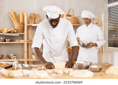 Professional African baker in white chef uniform kneading dough on flour-covered table in artisan bakery shop, surrounded by freshly baked bread and pastries arranged on wooden shelves - Powered by Shutterstock