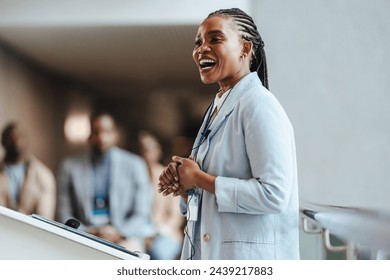 Professional African woman with a radiant smile engages attendees as she speaks at a business conference. With a blurred team in the backdrop, she embodies leadership and confidence. - Powered by Shutterstock