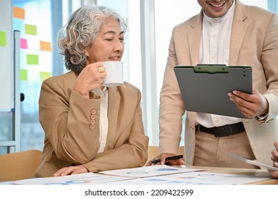 Professional Aged Asian Female Boss Talking And Giving Advice To Her Male Staff While Sipping Her Coffee During The Meeting.