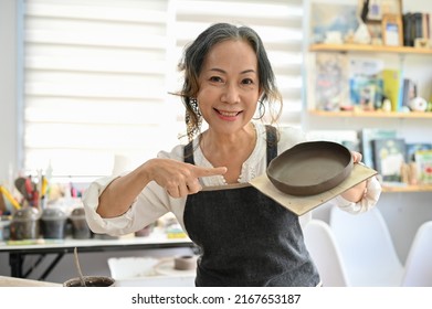 Professional Aged Asian Female Potter Showing Her Ceramic Plate In Her Studio. Beautiful Aged Asian Woman Joining A Pottery Workshop.