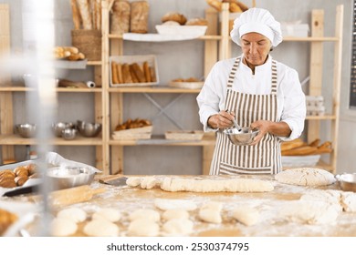 Professional aged female baker in white uniform and striped apron using whisk to mix ingredients in metal bowl, standing in small artisan bakery, surrounded by variety of freshly baked bread and tools - Powered by Shutterstock