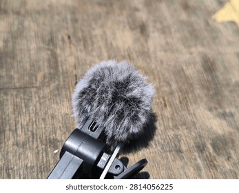 A professional, angled studio-style shot of a compact wireless microphone system resting on a dark, textured wooden surface. The focus is sharp on the device, showcasing its matte black finish, the dedicated power button, and the integrated clothing clip. A high-quality, grey synthetic fur windscreen is attached to the microphone to reduce noise during outdoor recording sessions. The composition uses natural light to create a clean, modern aesthetic that highlights the build quality and design details of the device. This image is ideal for use in technical reviews, e-commerce product pages, or educational content focused on audio engineering, portable recording setups, and gear for influencers, content creators, and videographers who demand reliable performance and aesthetic appeal in their professional audio and video production tools.