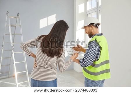 Professional architect, engineer or builder foreman in a yellow uniform and a white hard hat holding a house construction blueprint plan and talking to a young woman homeowner. Back view from behind