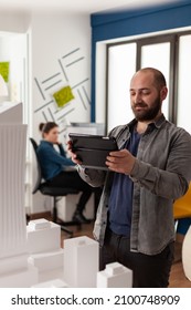 Professional Architect Holding Tablet Smiling In Videocall Conference Presenting Final Maquette To Client. Bearded Architectural Engineer Photographing Table With White Foam Residential Model.