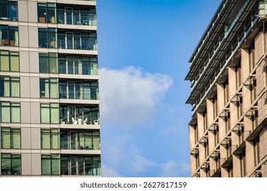 A professional architectural composition featuring two modern high-rise buildings facing each other against a clear blue sky with a few soft white clouds. On the left, a contemporary apartment complex displays a grid-like facade with large glass windows, sliding glass doors, and private balconies. Visible interior details include white curtains and furniture, suggesting a luxury residential living space. On the right, a contrasting commercial or office building exhibits a repetitive geometric pattern of vertical concrete pillars and recessed windows with integrated exterior sunshades or louvers. The image emphasizes the verticality and linear perspective of urban development. The lighting is bright and natural, highlighting the textures of glass, steel, and concrete. The composition creates a narrow vertical channel of sky between the two structures, illustrating dense urban planning and modern city architecture. High-angle perspective looking upwards, focusing on the intersection of residential and corporate real estate in a metropolitan environment. Clean lines, symmetrical elements, and a cool color palette dominated by blue and grey tones.