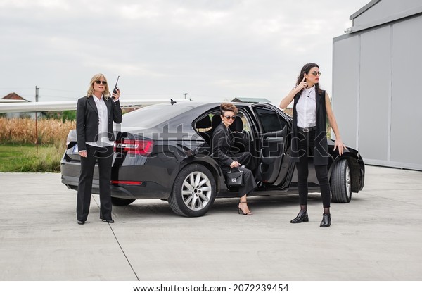Professional armed team of female and male\
bodyguards protect celebrity person in car limousine. Bodyguard and\
VIP person security protection. Professional police agent in\
civilian black\
suit
