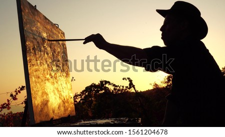 Professional artist paints a picture in nature. Silhouette of an adult man, sunset, abstract autumn landscape. Painting in the open air using a portable easel and a paintbox