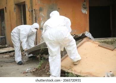 Professional asbestos removal. 
Worker with white protective suit, gloves and mask removes removal white asbestos on construction site. Demolition building, barrier tape warning, caution hazard. 