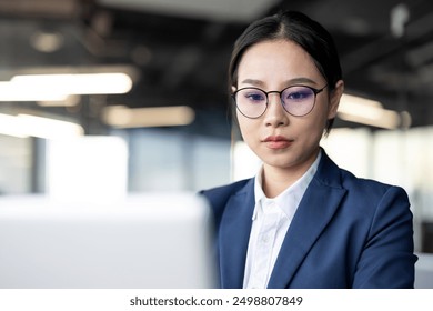 Professional asian businesswoman wearing glasses and formal attire, concentrating on her work while using a laptop in a contemporary office setting. The image depicts focus, productivity. - Powered by Shutterstock