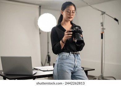 A professional Asian female photographer is focusing on adjusting her DSLR camera, working in a photoshoot studio with lighting equipment. - Powered by Shutterstock