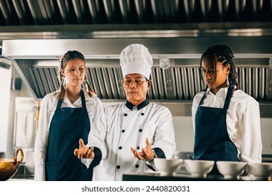 Professional Asian senior chef guides diverse students in a restaurant kitchen workshop focusing on teamwork learning and note-taking. Educational setting. Food Edocation - Powered by Shutterstock