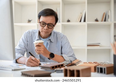 A professional Asian senior male interior designer or architect is sipping coffee while working on his project in the office. home construction business concept - Powered by Shutterstock