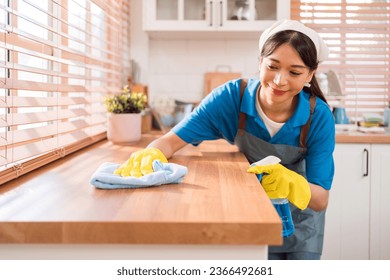 Professional Asian woman cleaning service wearing yellow rubber gloves, using a rag to wipe with spraying liquid detergent on the wooden top kitchen counter at home. Housekeeping cleanup, cleaner. - Shutterstock ID 2366492681