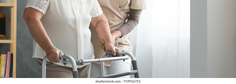 Professional assistant supporting elderly woman in white shirt using walker in living room
