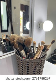 Professional assorted makeup brush set in a stylish woven holder by a lighted vanity mirror. Perfect for beauty tutorials, makeup artists, and vanity decor.