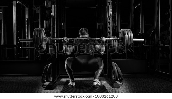 Professional athlete sits\
with a barbell on his shoulders and prepares to stand with her.\
View from the\
back.