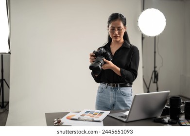 A professional, attractive Asian female photographer is adjusting her DSLR camera for a photoshoot, working in a professional studio with lighting equipment. - Powered by Shutterstock