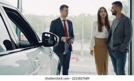 Professional Automobile Seller In Suit Is Having Conversation With Attractive Rich Couple Telling Them About New Car Model, Cheerful Buyers Are Smiling. Focus On Beautiful Shiny Auto.