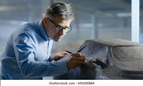 Professional Automotive Designer With Rake Sculpts Futuristic  Car Model From Plasticine Clay. He Works In A Special Studio Located In A Large Car Factory.